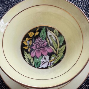 May include: A vintage teacup with a floral design. The cup is white with a gold rim and features a pink and yellow flower with green leaves on a black background.