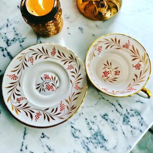 Pu&ograve; includere: Un set di tazza e piattino da t&egrave; vintage. La tazza e il piattino in porcellana bianca sono adornati con finiture dorate e un motivo di foglie marroni e bacche rosse. La tazza ha un manico dorato. Il set &egrave; esposto su una superficie di marmo.