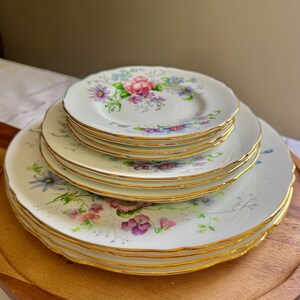 May include: A collection of vintage white porcelain plates featuring a floral pattern in pink, purple, and blue hues. Each plate has a gold rim and a scalloped edge. The plates are stacked on a wooden surface, showcasing a classic design.