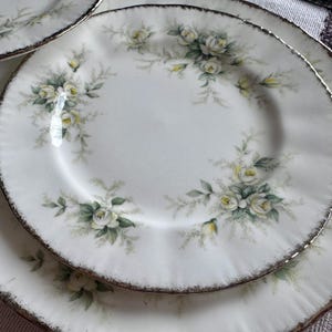 May include: Two white ceramic plates with a floral design featuring white roses and green leaves. The plates have a gold rim.