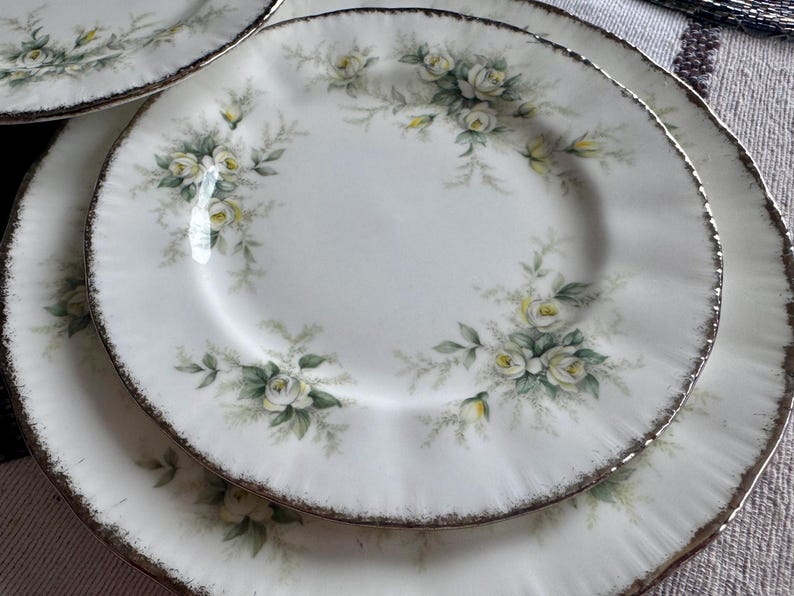 May include: Two white ceramic plates with a floral design featuring white roses and green leaves. The plates have a gold rim.