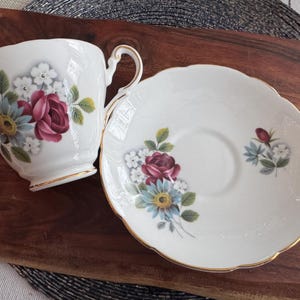 May include: A white teacup and saucer set with a floral design featuring pink and red roses, blue and white flowers, and green leaves.