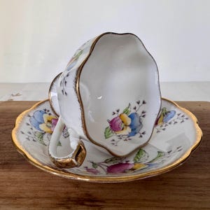 May include: White teacup and saucer set with a floral design in blue, yellow, and purple. The edges of the cup and saucer are scalloped and trimmed with gold. The teacup is resting on the saucer.