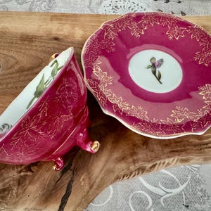 May include: A vintage teacup and saucer set. The cup and saucer are a deep pink with gold leaf accents. The inside of the cup is white with a floral design. The saucer has a floral design in the center.