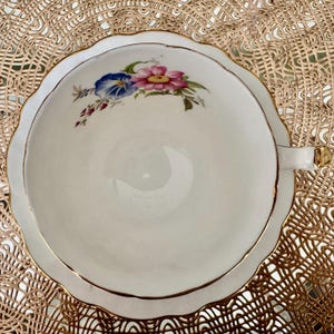 May include: A white teacup with a gold rim and a floral design featuring pink, blue, and green flowers. The teacup is on a gold lace doily.