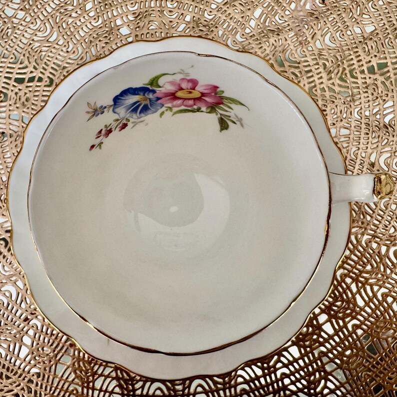May include: A white teacup with a gold rim and a floral design featuring pink, blue, and green flowers. The teacup is on a gold lace doily.