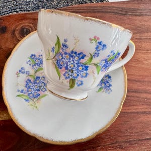 May include: A white teacup and saucer with a gold rim. The teacup and saucer are decorated with a floral pattern of blue forget-me-not flowers.