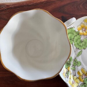 May include: A white teacup with a gold rim and a matching saucer with a floral design featuring yellow flowers and green leaves.