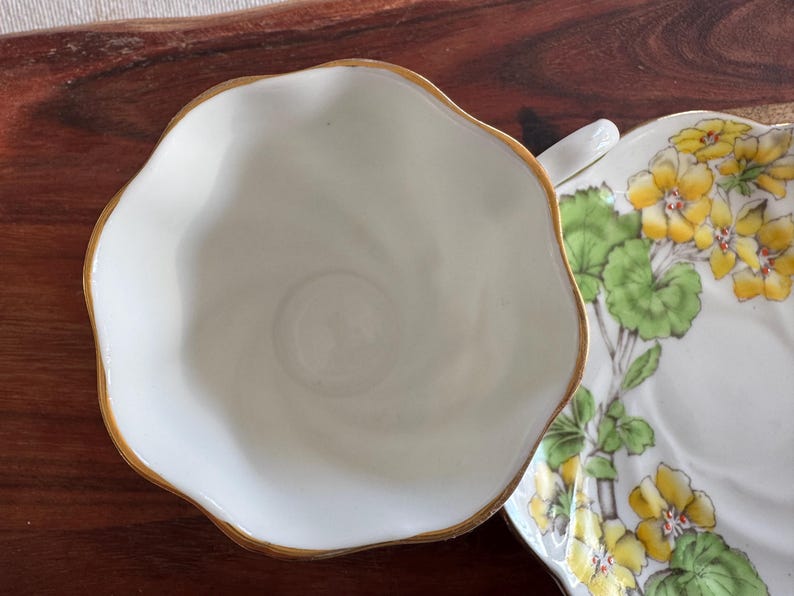 May include: A white teacup with a gold rim and a matching saucer with a floral design featuring yellow flowers and green leaves.
