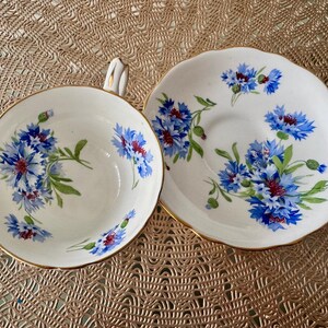 May include: A white teacup and saucer with a blue and white floral pattern. The floral pattern features blue cornflowers with green leaves and stems.