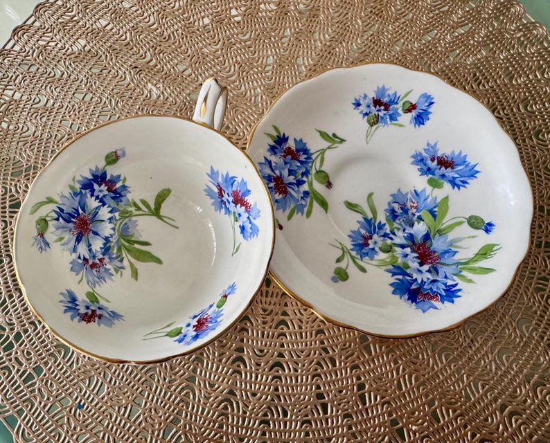May include: A white teacup and saucer with a blue and white floral pattern. The floral pattern features blue cornflowers with green leaves and stems.