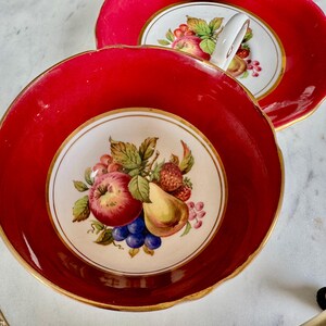 May include: A red teacup and saucer with a gold rim. The teacup has a white interior with a hand-painted design of fruit, including a pear, apple, grapes, and strawberries.