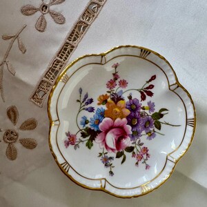 May include: A white, scalloped-edge porcelain dish with a floral bouquet design. The bouquet features pink, purple, blue, and yellow flowers. The rim is trimmed with gold. A decorative piece for serving or display.