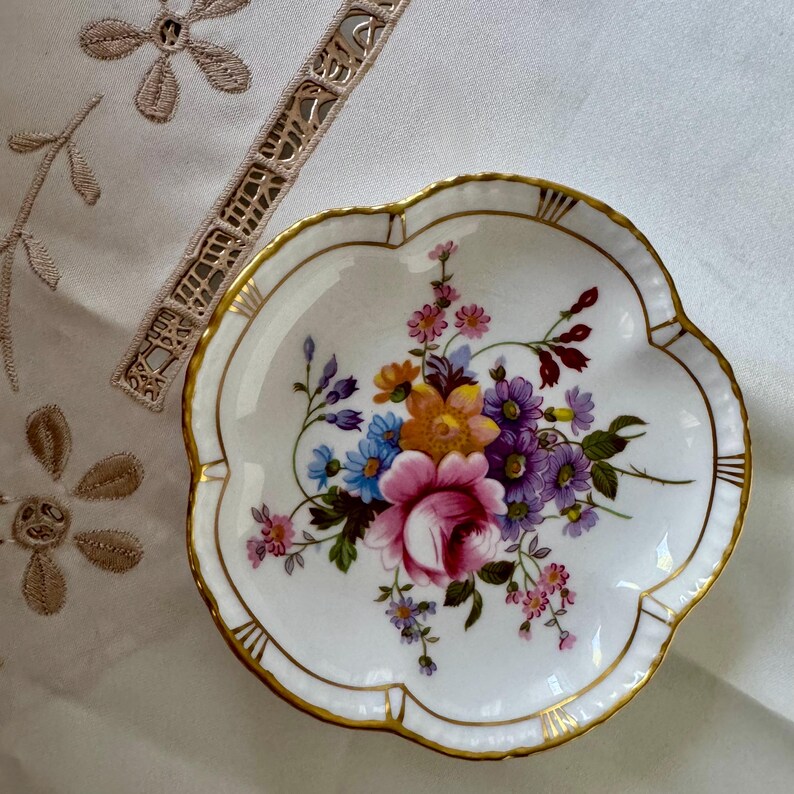 May include: A white, scalloped-edge porcelain dish with a floral bouquet design. The bouquet features pink, purple, blue, and yellow flowers. The rim is trimmed with gold. A decorative piece for serving or display.