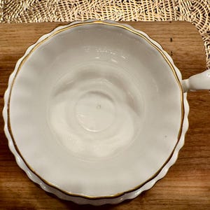 May include: White porcelain teacup with a gold rim and a delicate scalloped edge. The cup is empty and sits on a wooden surface.