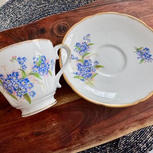 May include: A white teacup and saucer with a floral design featuring blue forget-me-not flowers. The cup and saucer have a gold trim.