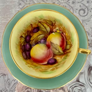May include: A vintage teacup and saucer set. The teacup interior features a hand-painted still life of peaches and grapes. The cup and saucer are a soft green with gold trim. The teacup handle is gold.