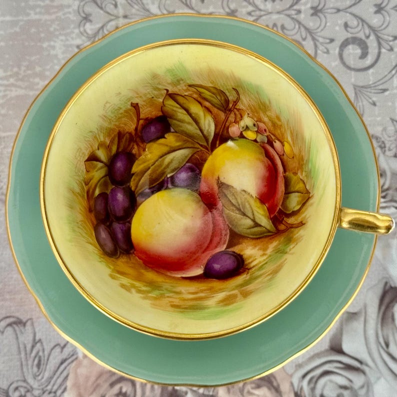 May include: A vintage teacup and saucer set. The teacup interior features a hand-painted still life of peaches and grapes. The cup and saucer are a soft green with gold trim. The teacup handle is gold.