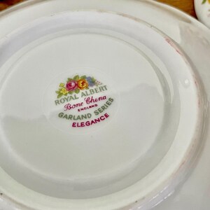 May include: Close-up of a white Royal Albert Bone China saucer from the Garland Series. The saucer features a floral emblem and the word "Elegance" in red. The saucer is part of a vintage tea set.