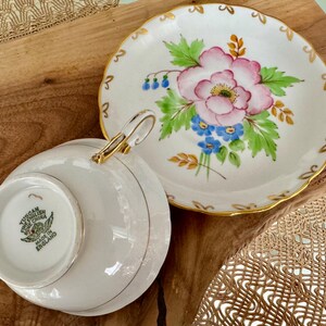 May include: A white teacup and saucer with a floral design. The saucer has a gold rim and a pink flower with blue and green leaves. The teacup has a gold rim and the words "Tucan English China Made in England" on the bottom.