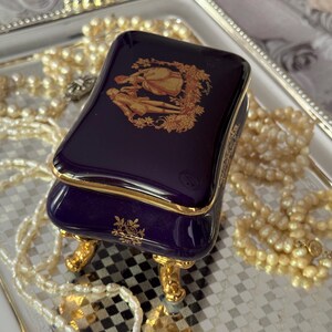 May include: A small, rectangular, navy blue jewelry box with gold trim and ornate gold feet. The lid features a gold illustration of two figures. The box is on a silver tray, surrounded by strands of pearls.