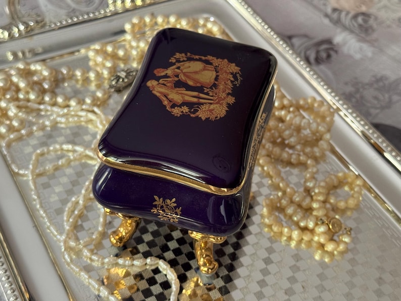 May include: A small, rectangular, navy blue jewelry box with gold trim and ornate gold feet. The lid features a gold illustration of two figures. The box is on a silver tray, surrounded by strands of pearls.
