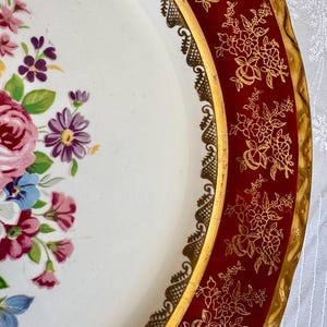 May include: A vintage decorative serving tray with a white center featuring a floral arrangement in pink, purple, and blue. The tray has a red border with gold floral designs and a gold trim. The tray's edge has a decorative gold lace design.