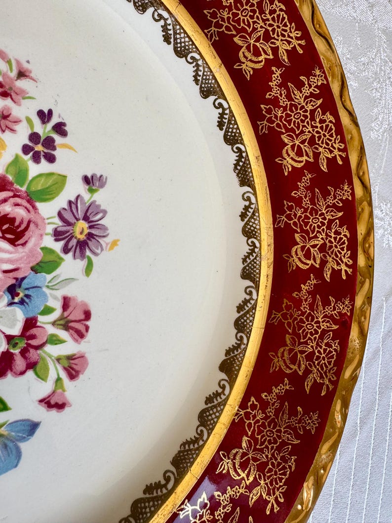 May include: A vintage decorative serving tray with a white center featuring a floral arrangement in pink, purple, and blue. The tray has a red border with gold floral designs and a gold trim. The tray's edge has a decorative gold lace design.