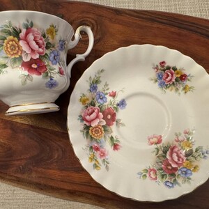 May include: A white teacup and saucer with a floral pattern. The cup and saucer have a gold trim. The flowers are pink, red, yellow, and blue.