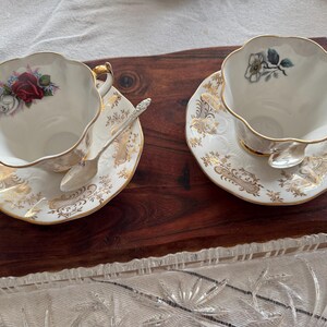 May include: Two white teacups with gold trim and floral designs on the inside. The teacups are on matching saucers with gold trim and floral designs. The teacups and saucers are on a wooden surface.