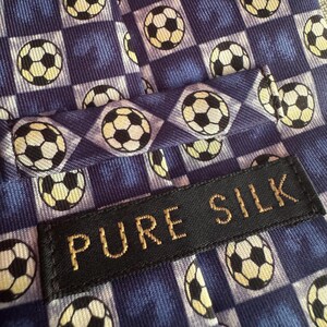 May include: A blue necktie featuring a repeating pattern of black and white soccer balls arranged in a checkered grid. A black rectangular fabric label is stitched onto the front, displaying the text PURE SILK in gold-colored embroidered letters.