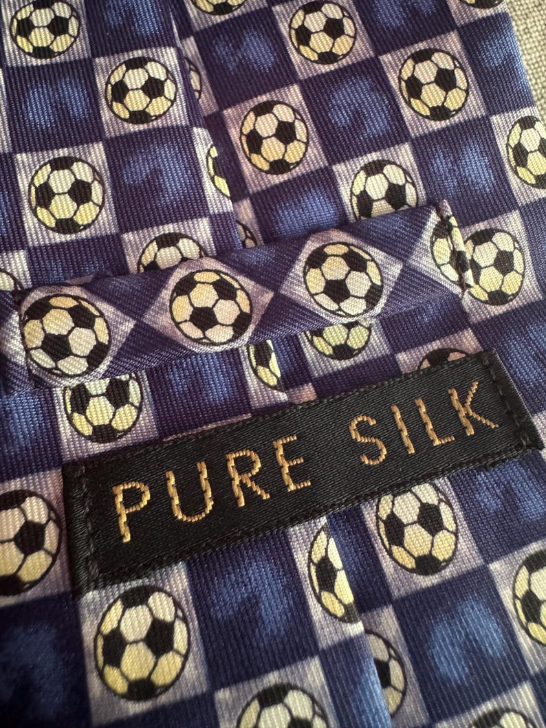 May include: A blue necktie featuring a repeating pattern of black and white soccer balls arranged in a checkered grid. A black rectangular fabric label is stitched onto the front, displaying the text PURE SILK in gold-colored embroidered letters.
