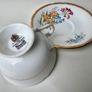 May include: A white teacup and saucer with a floral pattern. The saucer has a gold rim. The teacup has a gold band around the rim and a Paragon Fine Bone China England Reg'd mark on the bottom.