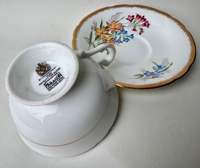 May include: A white teacup and saucer with a floral pattern. The saucer has a gold rim. The teacup has a gold band around the rim and a Paragon Fine Bone China England Reg'd mark on the bottom.