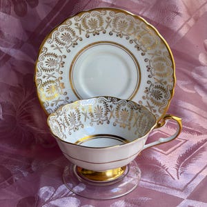 May include: A white porcelain teacup and matching saucer set featuring an intricate gold filigree pattern. The cup has a scalloped rim, a gold-trimmed base, and a curved handle. The saucer displays a matching ornate gold design along the edge.
