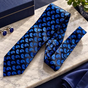 May include: A black necktie featuring a repeating blue paisley pattern, accompanied by a pair of round silver-toned cufflinks with dark blue circular insets. The necktie has a smooth, satin-like finish and a pointed end, displayed alongside the cufflinks.