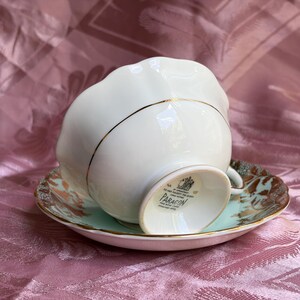 May include: A cream-colored porcelain teacup and saucer set with a scalloped edge and gold trim. The underside of the cup features a black stamp with the text BY APPOINTMENT TO HER MAJESTY THE QUEEN PARAGON FINE BONE CHINA MADE IN ENGLAND.