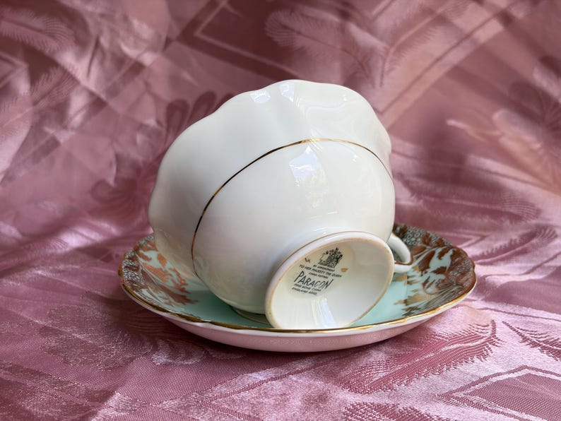 May include: A cream-colored porcelain teacup and saucer set with a scalloped edge and gold trim. The underside of the cup features a black stamp with the text BY APPOINTMENT TO HER MAJESTY THE QUEEN PARAGON FINE BONE CHINA MADE IN ENGLAND.