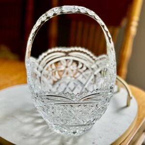 May include: A clear, cut-glass basket with a decorative handle. The basket features intricate floral and geometric patterns throughout its design. The handle is also cut with a decorative pattern. The basket is sitting on a white surface.