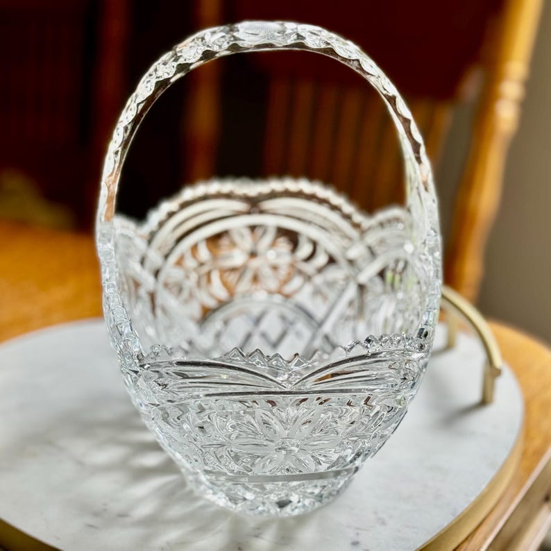 May include: A clear, cut-glass basket with a decorative handle. The basket features intricate floral and geometric patterns throughout its design. The handle is also cut with a decorative pattern. The basket is sitting on a white surface.