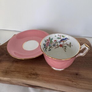 May include: A pink teacup and saucer set with gold trim. The teacup features a detailed illustration of a blue bird perched on a flowering branch. The saucer is pink with a white center.