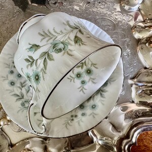 May include: A white teacup and saucer set with a floral pattern in shades of blue and green, accented with silver trim. The teacup is resting on its side on a silver tray, with a pearl necklace in the background.