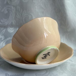 May include: A peach-colored teacup and saucer set with a scalloped edge. The underside of the teacup features a green rim and a stamp that reads Pastella Royal Albert Bone China England. The interior of the cup and the saucer are white.