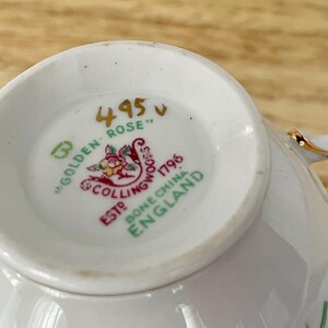 Puede incluir: Primer plano del interior de una taza de t&eacute; blanca, que muestra el dise&ntilde;o "Golden Rose". La base presenta un emblema detallado con elementos florales y texto, incluyendo "Bone China England". La taza tiene ribete dorado.