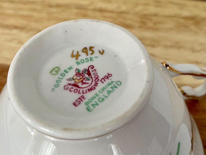 Puede incluir: Primer plano del interior de una taza de t&eacute; blanca, que muestra el dise&ntilde;o "Golden Rose". La base presenta un emblema detallado con elementos florales y texto, incluyendo "Bone China England". La taza tiene ribete dorado.