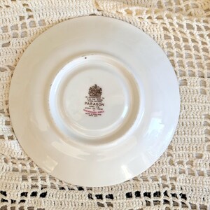 May include: A white ceramic saucer with a circular indentation in the center. The underside features a brown and red stamp that reads By Appointment to H.M. The Queen China Potters Paragon China Ltd. Stoke-on-Trent Marlborough Series Made in England.