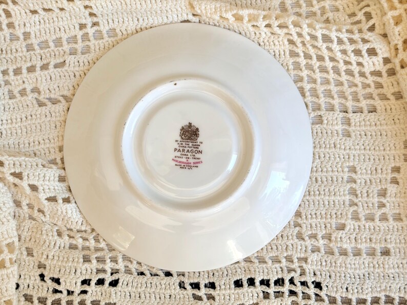 May include: A white ceramic saucer with a circular indentation in the center. The underside features a brown and red stamp that reads By Appointment to H.M. The Queen China Potters Paragon China Ltd. Stoke-on-Trent Marlborough Series Made in England.