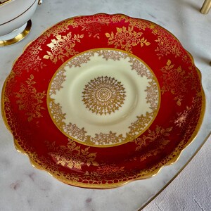 May include: A red and gold decorative plate and saucer set. The plate has a scalloped edge and is red with gold floral designs. The saucer is cream-colored with a gold floral design in the center and around the edge. The set is likely vintage.