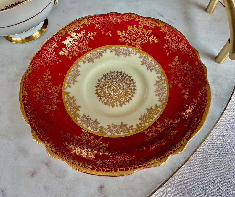 May include: A red and gold decorative plate and saucer set. The plate has a scalloped edge and is red with gold floral designs. The saucer is cream-colored with a gold floral design in the center and around the edge. The set is likely vintage.
