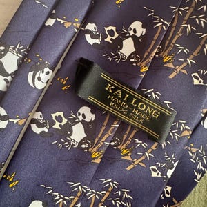May include: A dark blue necktie featuring a repeating pattern of pandas eating bamboo stalks. A black fabric label is stitched onto the front, displaying the gold text KAI LONG, HAND-MADE, and 100% SILK in a clear, legible font.